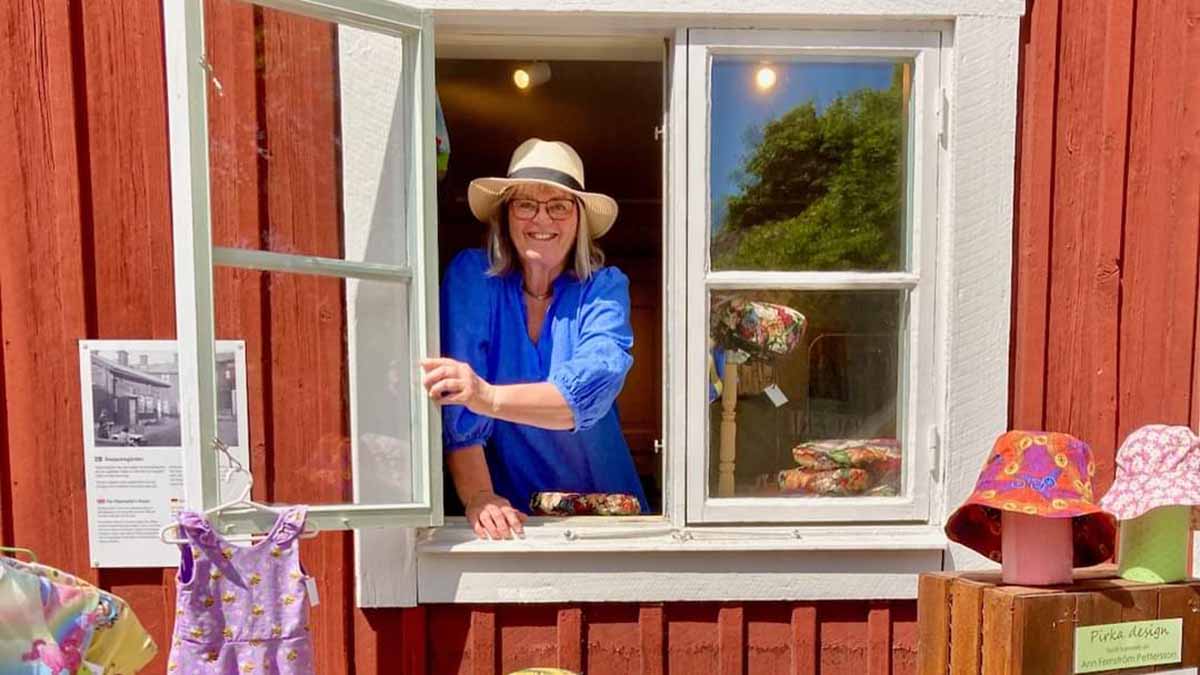 Glad person tittar ut genom ett öppet fönster mot faluröd bakgrund. Framför fönstret hänger färgglada kläder. 