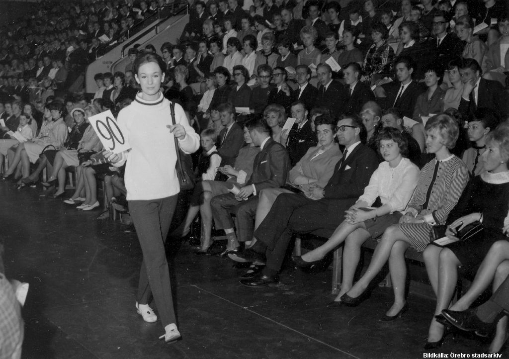 En ung kvinna i ljus tröja och mörka byxor går framför publik.
