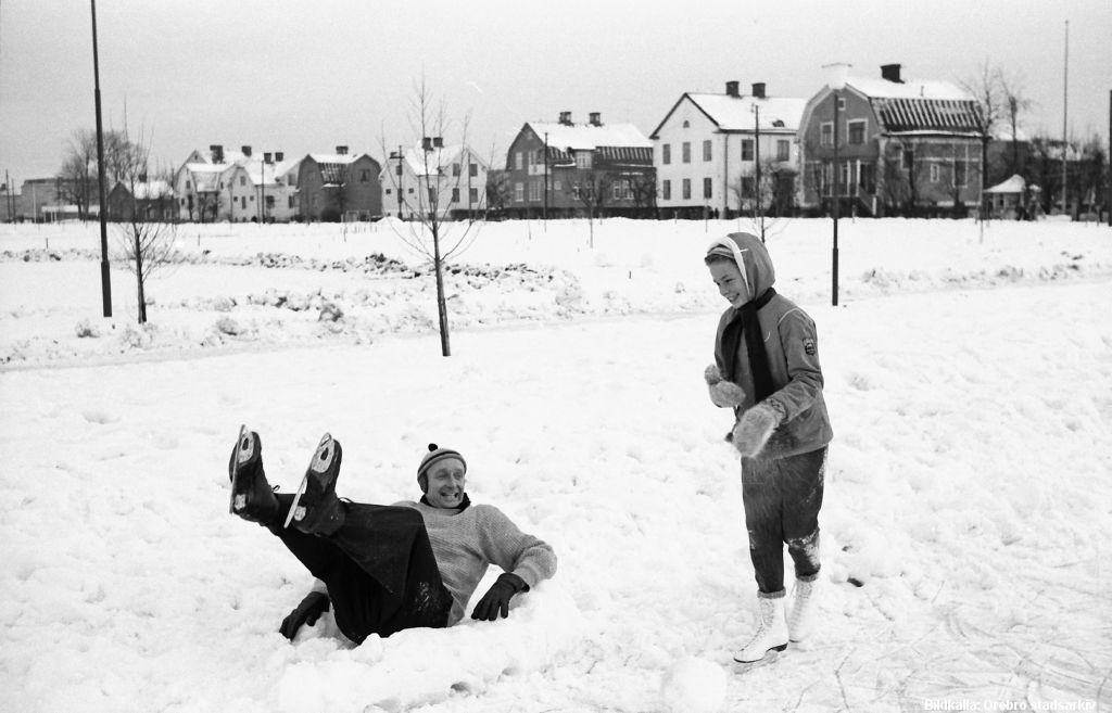 En man ligger i snödriva med skridskoklädda fötter i luften. En kvinna på skridskor står brevid och ler.