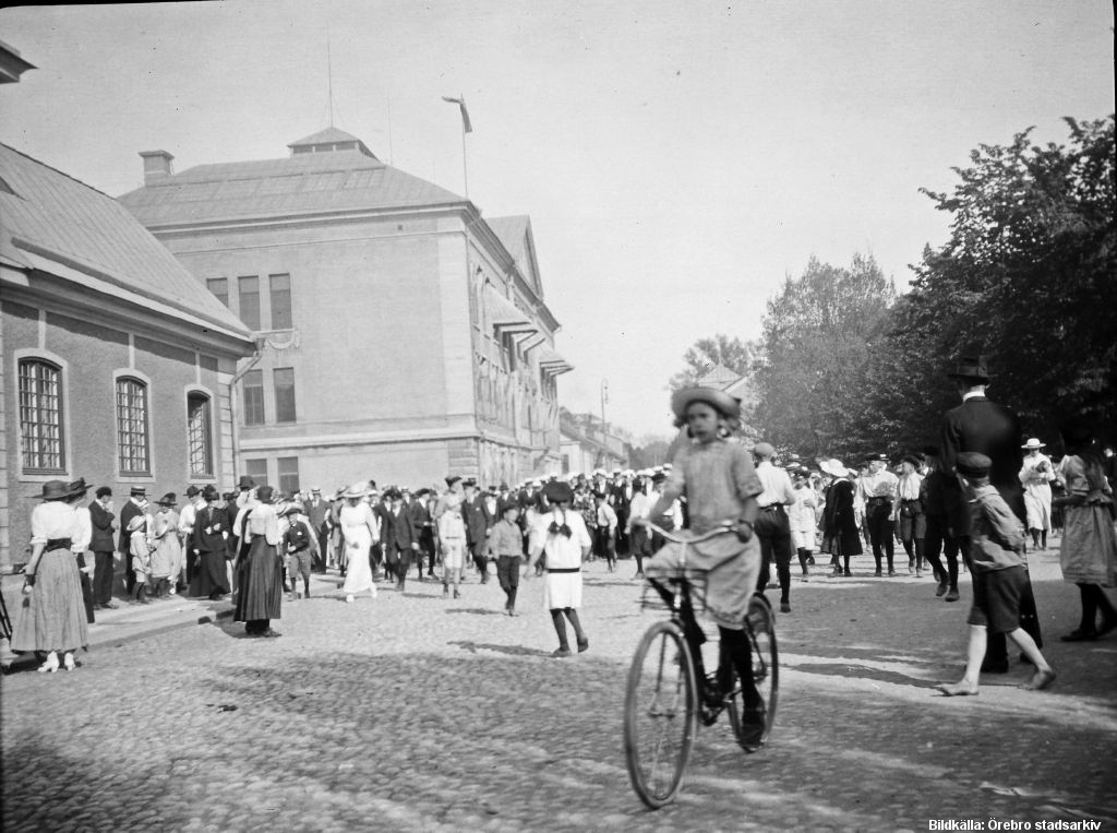 Folkmassa samlad framför stort stenhus (Karolinska skolan). En flicka cyklar längst fram mot fotografen.
