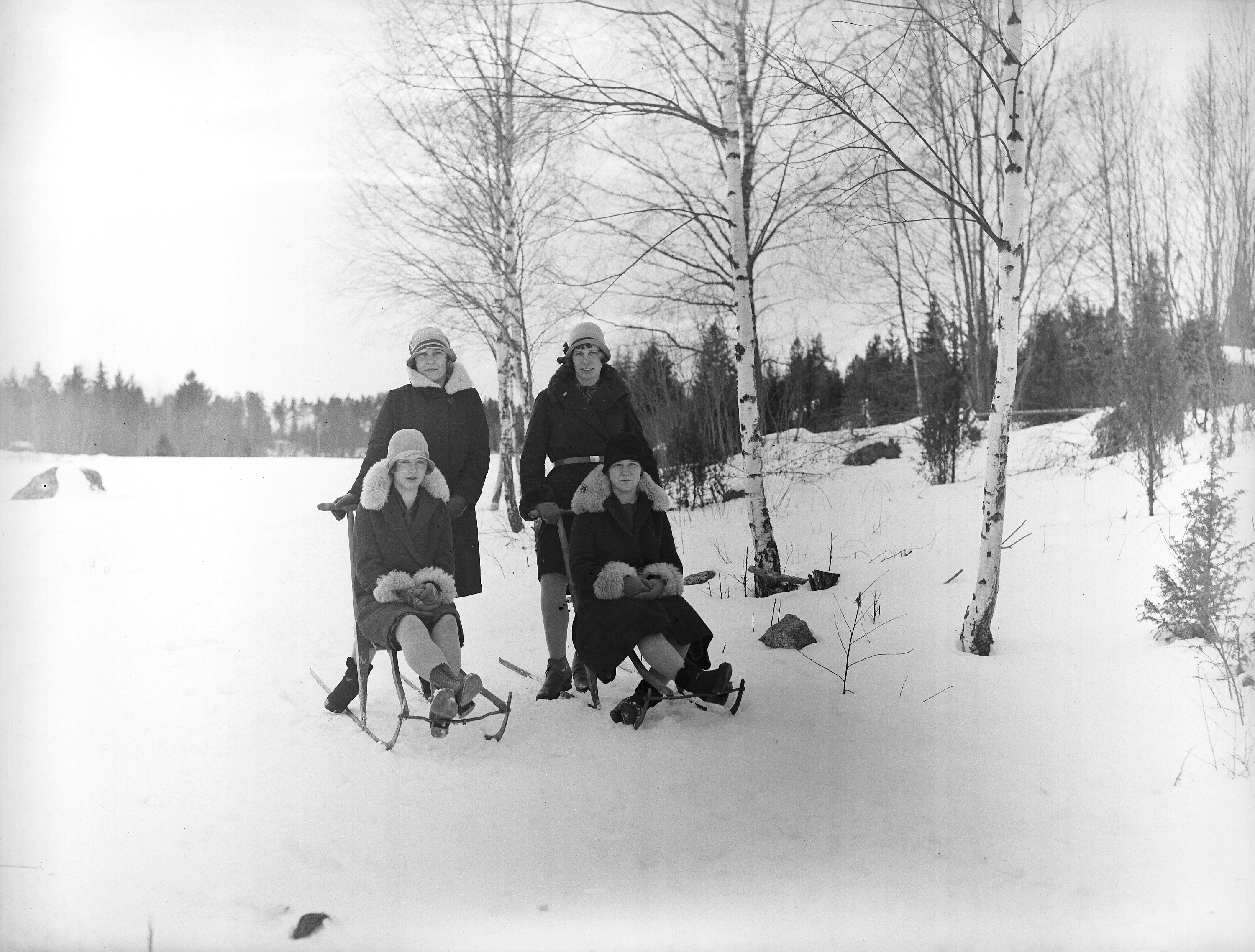 Två kvinnor står bakom två andra kvinnor som sitter på varsin spark, i snötäckt landskap.