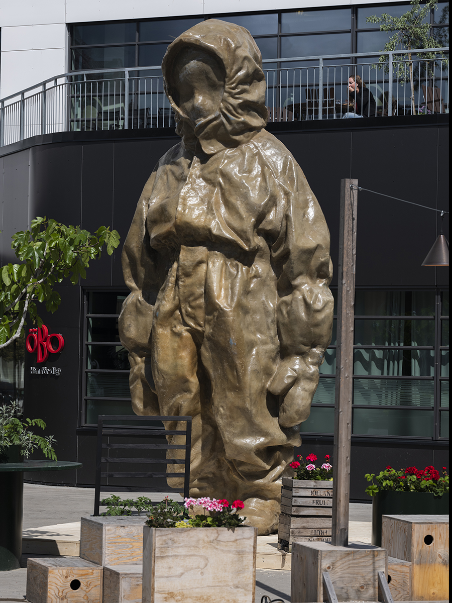 I en stadsmiljö står en fem meter lång brungul staty, det föreställer kanske ett barn i en overall med luvan uppdragen