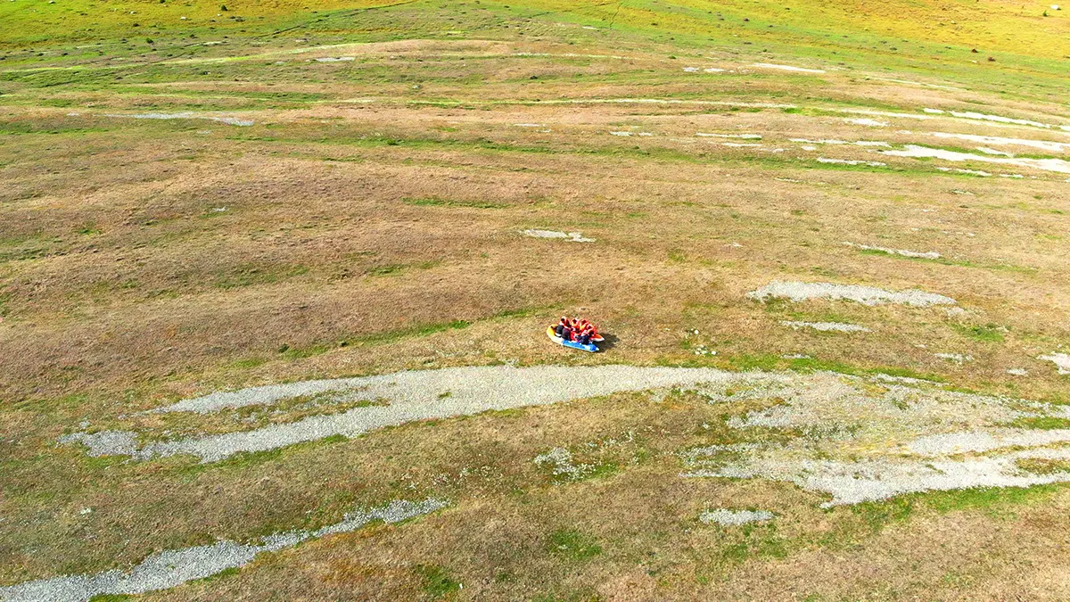 En bild ifrån videoverket The Flood. Bilden består av ett landskap där marken är grön och i mitten ser man människor sittandes i en gummibåt.
