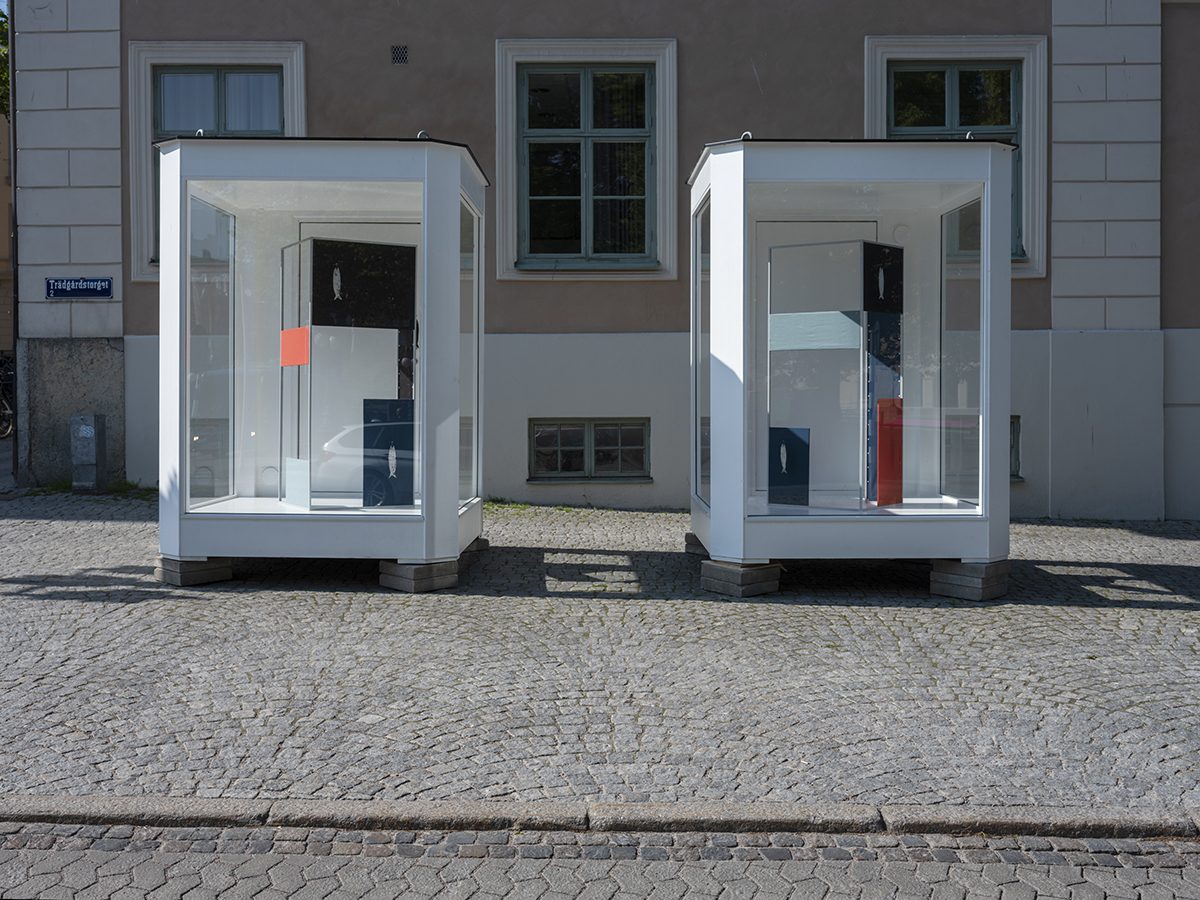 Två moduler med fönsterglas och vita karmar är placerade på en gata med kullersten. Inuti modulerna syns skulpturer.