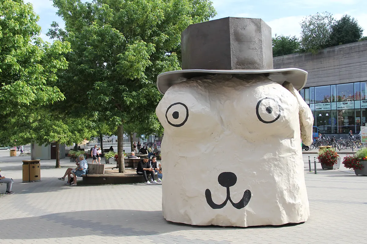 På ett torg står en beige byggnad formad som ett djurhuvud med hatt. Huvudet har två ögon och en nos.