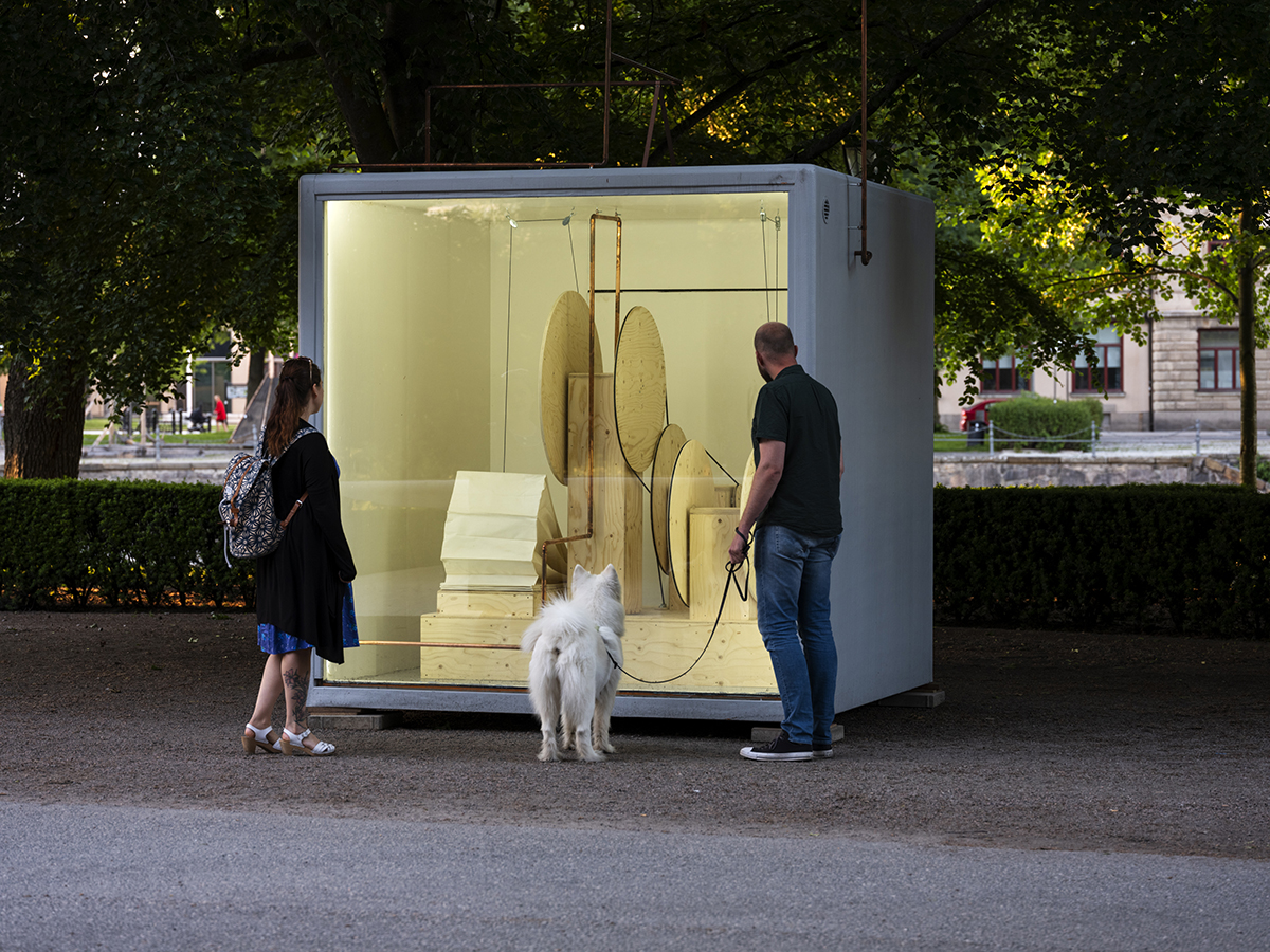 Två personer tittar in i en vit kub med glasfönster som är placerad i en park. Inuti kuben står en trären maskin med runda delar, blåsbälg och rör.
