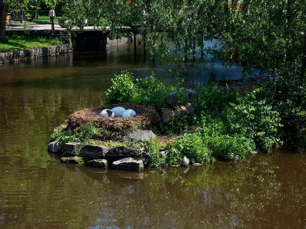 På en liten ö i en å finns en installation i form av ett stort fågelbo med tre ägg i stenmaterial. Ett av äggen ser ut att ha kläckts och är tomt medan de andra två är hela. Installationen omges av olika växter och skuggas av ett träd. I bakgrunden syns en bro och en park.