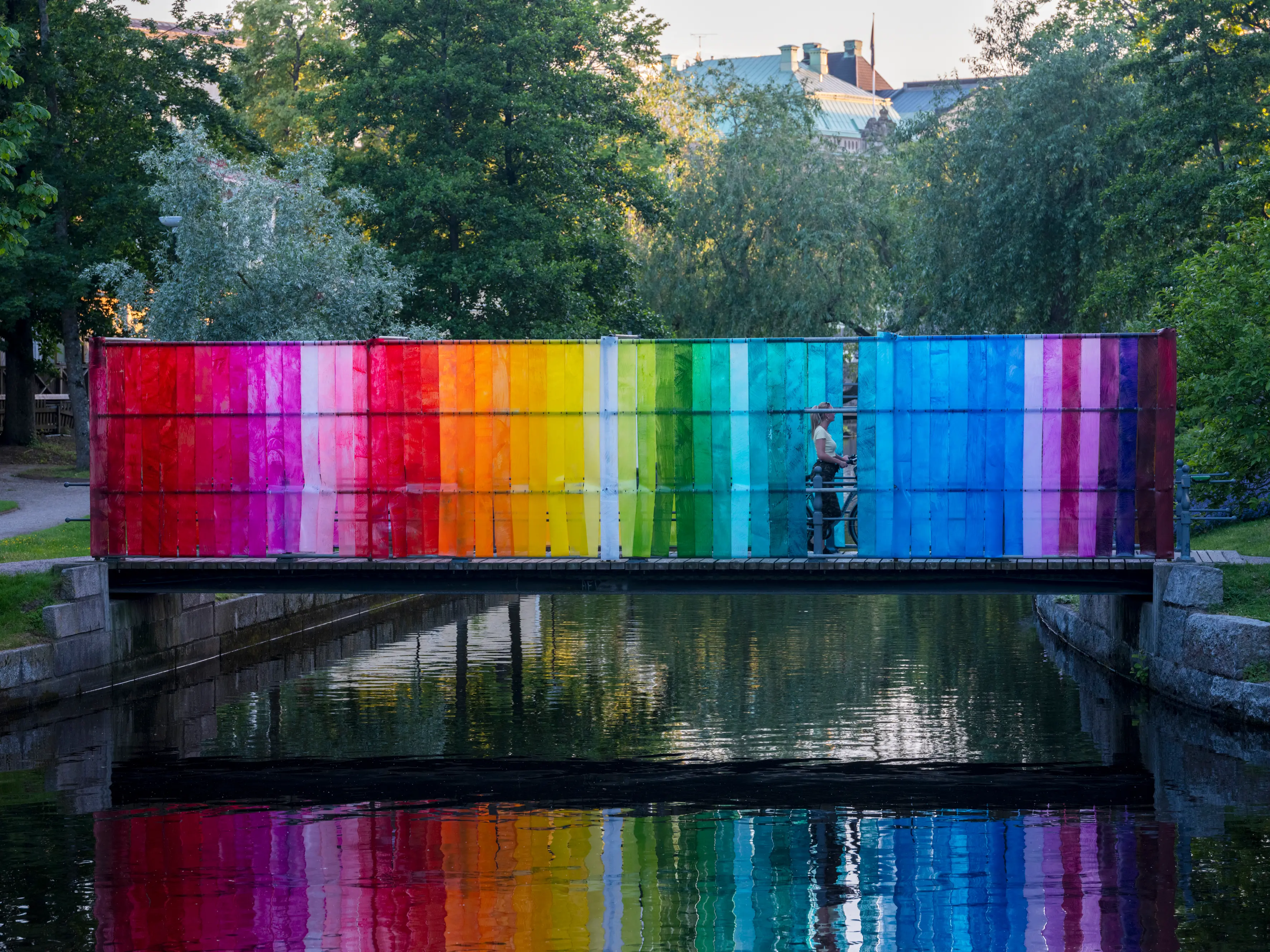 En person på cykel har stannat på bro med regnbågens alla färger som speglar sig i vattnet nedanför. 