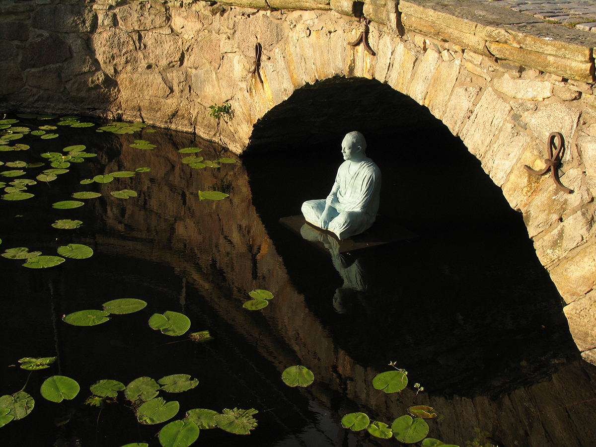Under en bro sitter en skulptur av en man som mediterar på vattenytan. 