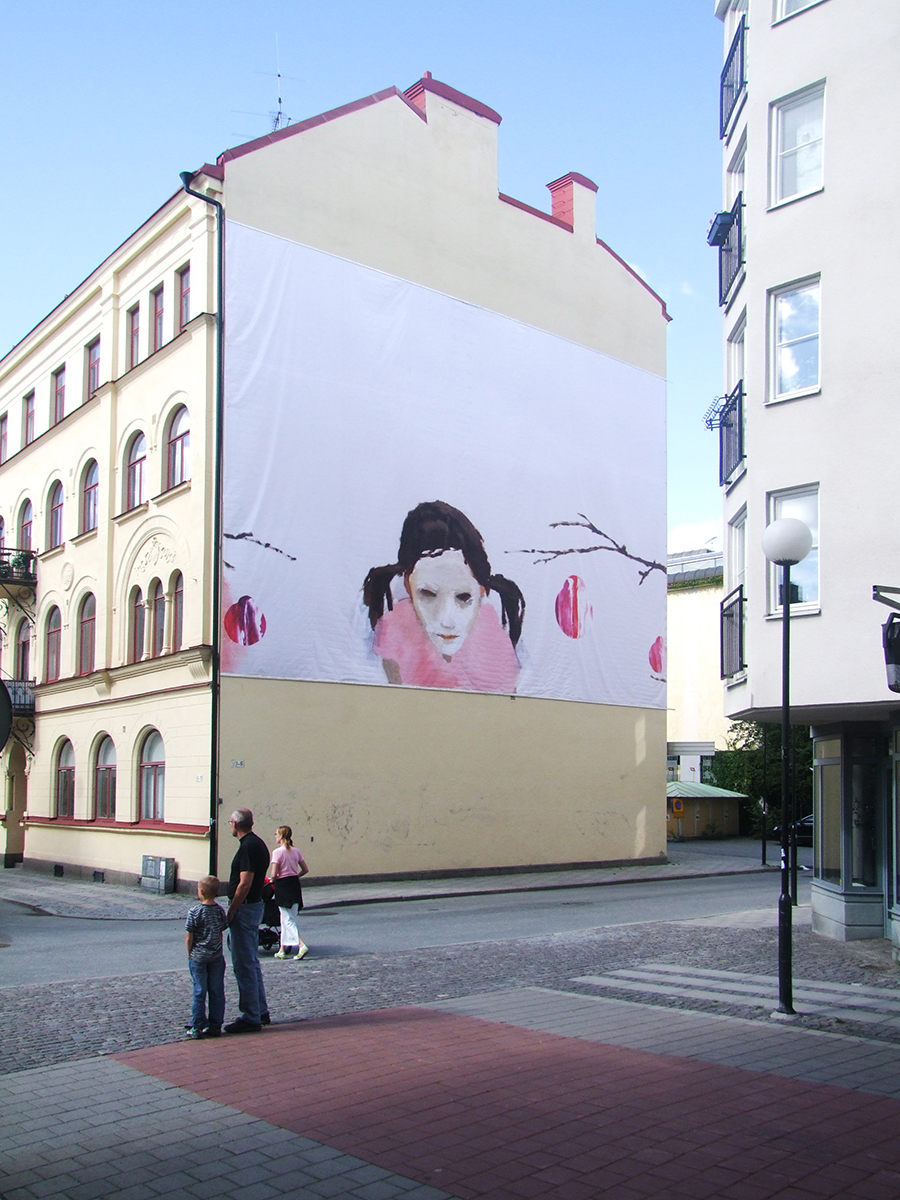 På en gul husfasad hänger en vepa som föreställer en målning av en flicka med rosa T-shirt och två tofsar. 