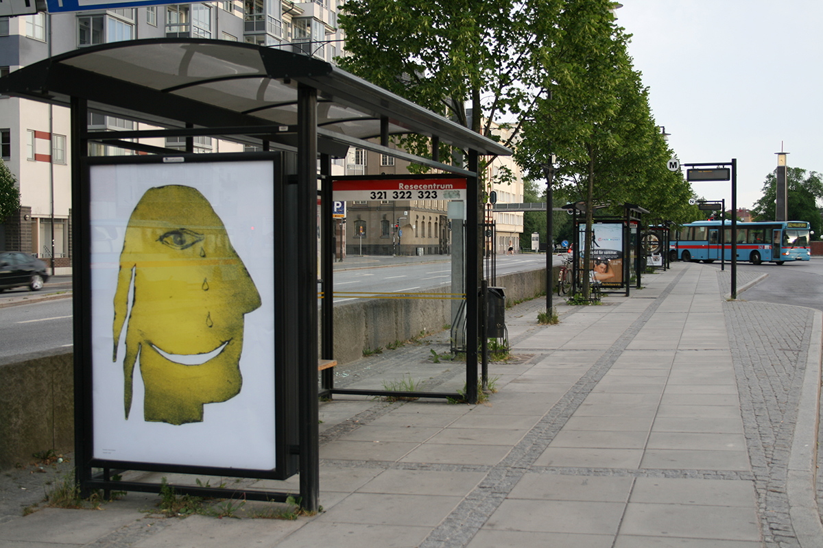 På ett busshållplats skyltfönster sitter en vit affisch. På affischen är en målning av en ett gult ansikte som fäller en tår. 