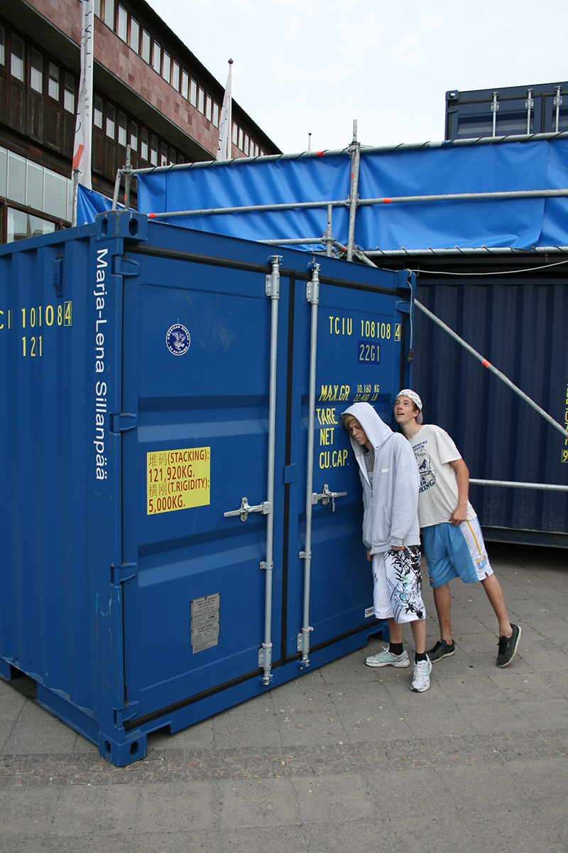 Två killar står och hänger emot en blå container. 