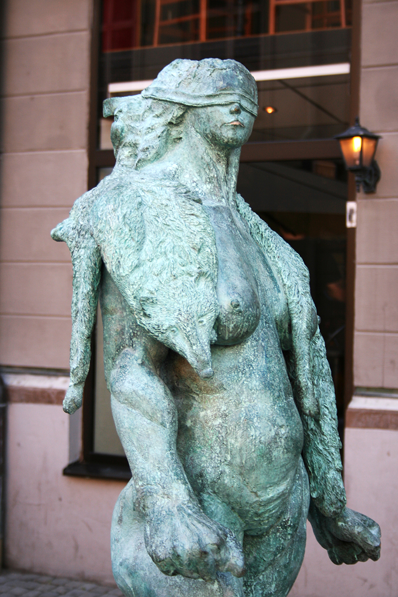 På torget står en staty av en naken kvinna som har en rävpäls runt sin nacke och hennes ögon är täckte med ett band. 
