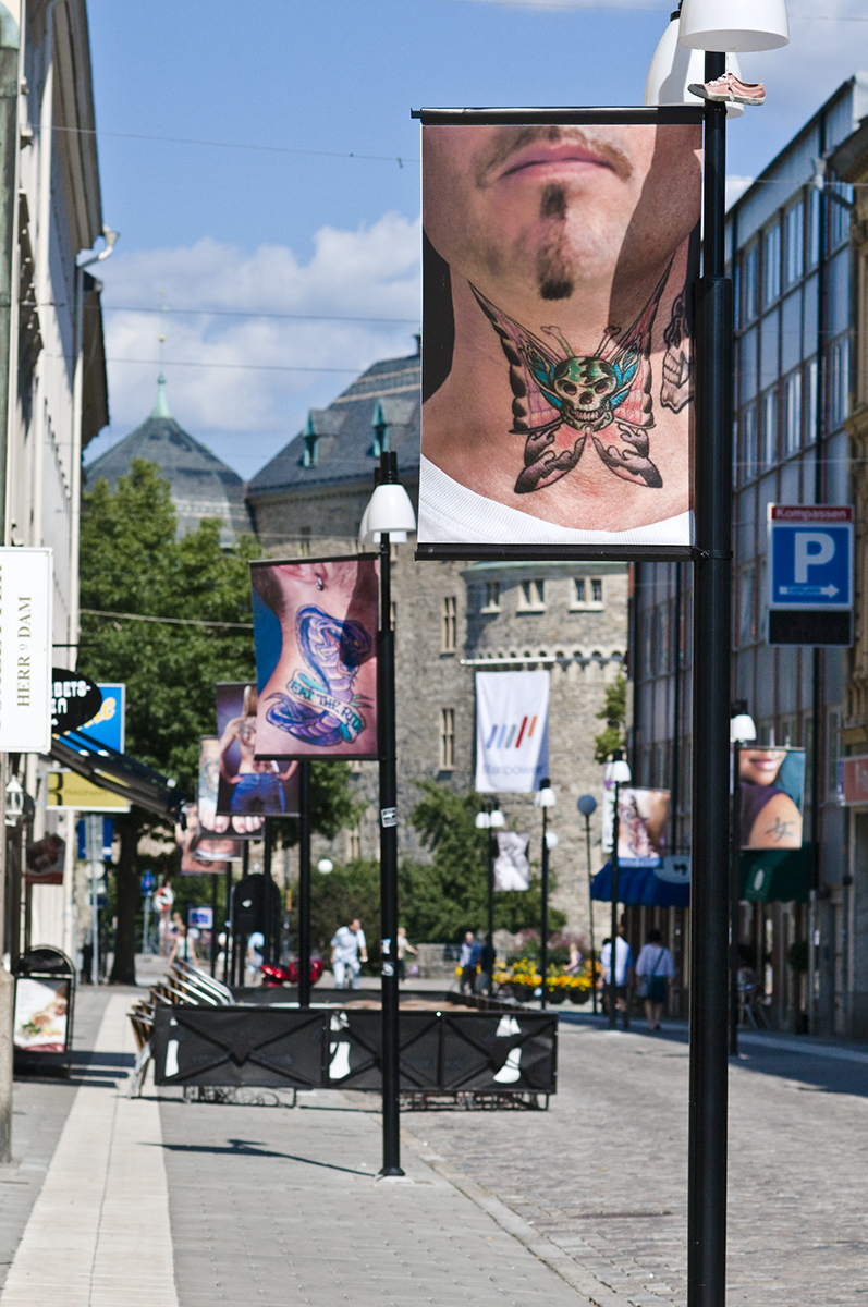 På alla lyktstolpar längs en gata hänger vepor föreställandes fotografier på folks tatueringar. 