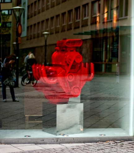 I ett glasfönster står en röd staty liknande en stor bil motor på en vit piedestal. 