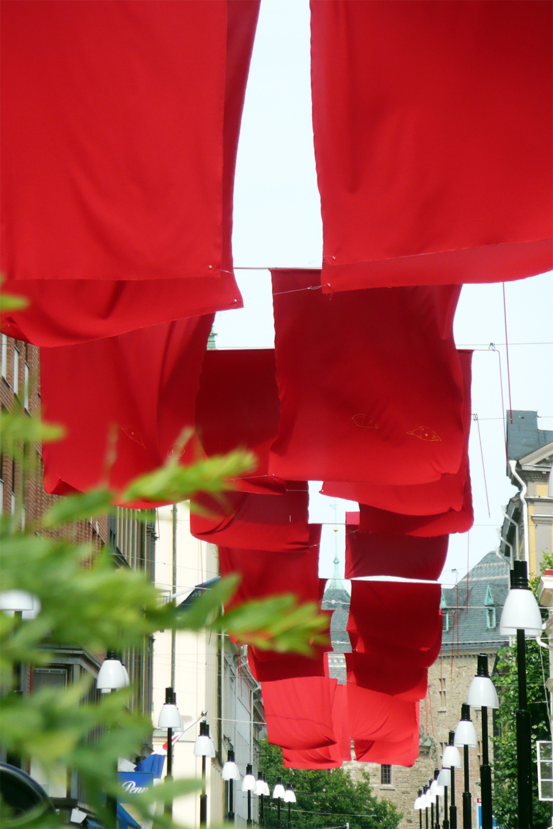 Mellan husfasader är rep fästa och på dessa hänger röda flaggor ner från luften som följer en gata.