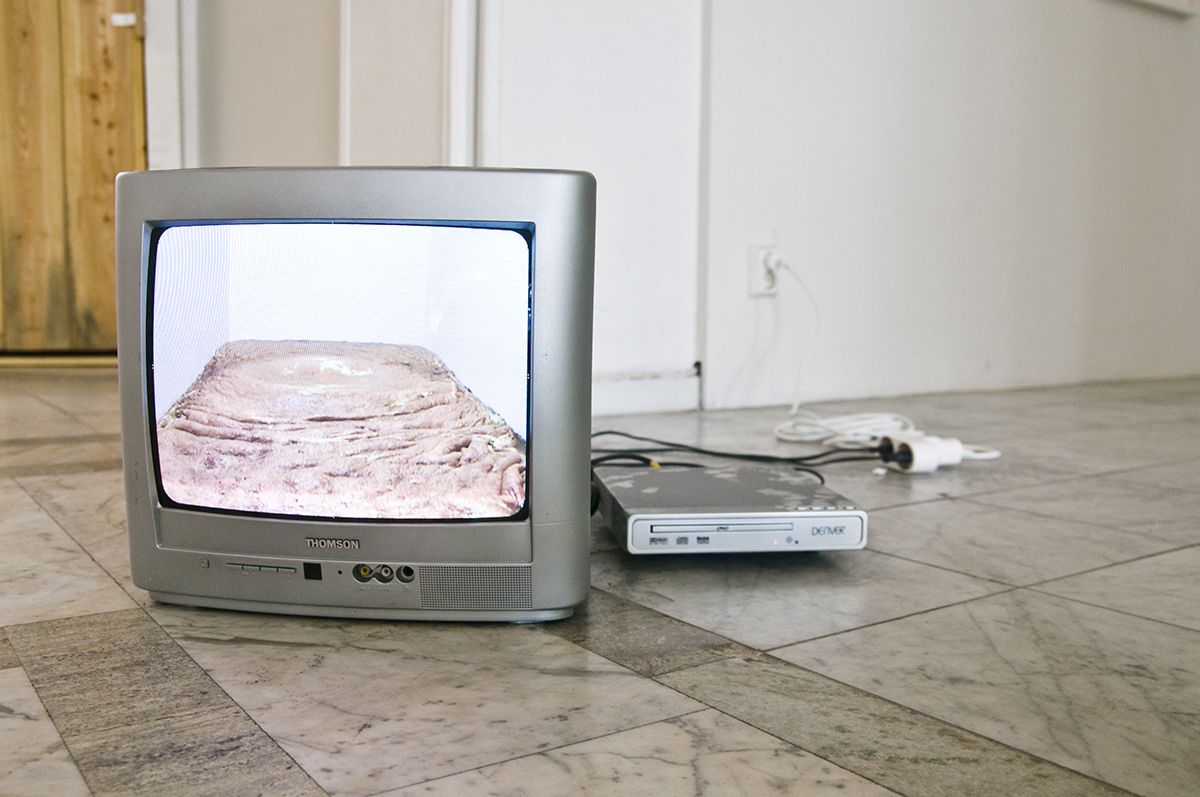 På ett golv står en silverfärgad analog TV samt en separat DVD-spelare. TV skärmen visar ett vitt rum med en orangefärgad matta som täcker hela golvet.