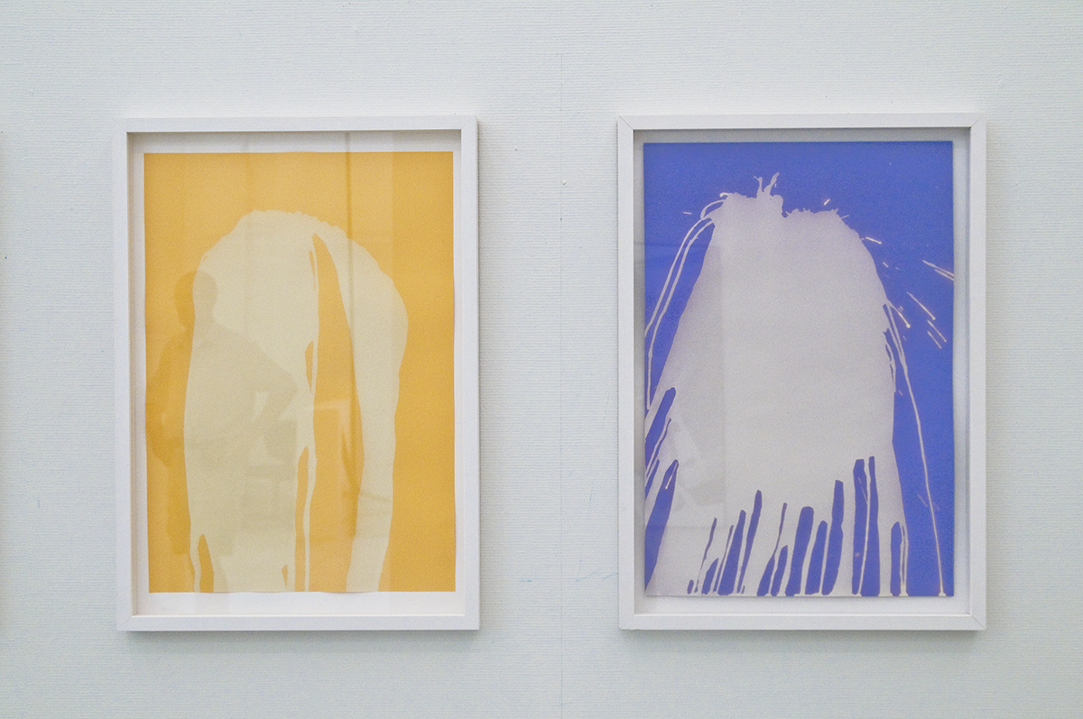 På en vit vägg hänger två inramade målningar. En med blå bakgrund och en med gul sedan ser det ut som att konstnären har kastat vit färg på pappret.