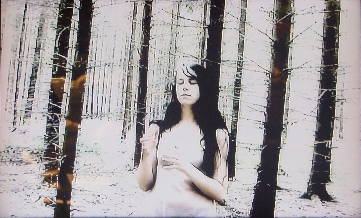 En bild ur en videosekvens där en kvinna iklädd vitt står i en skog.