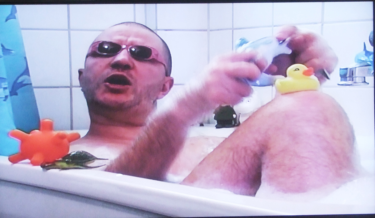 På bilden sitter en man och badar i ett badkar, han har lite försmå solbrillor på sig och omringas av badleksaker.