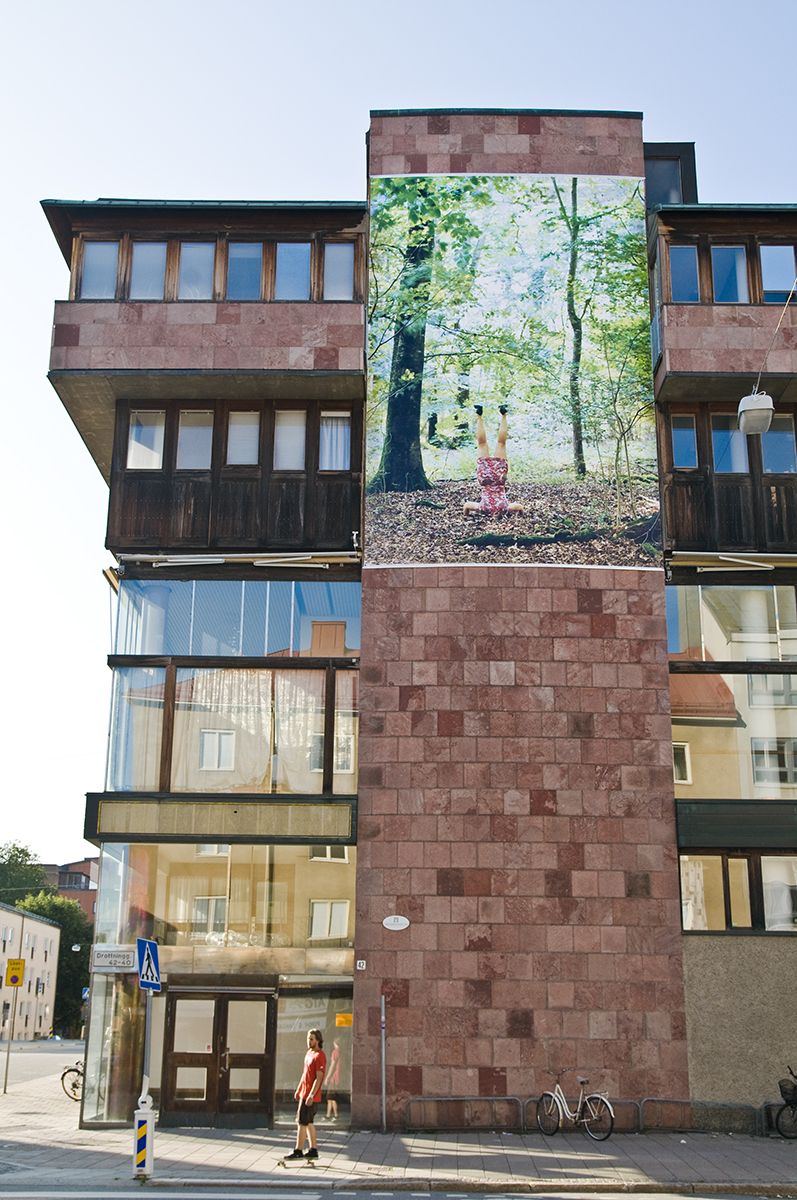 På en fasad sitter en vepa föreställandes ett fotografi av en kvinna som står på huvud i en skog. 