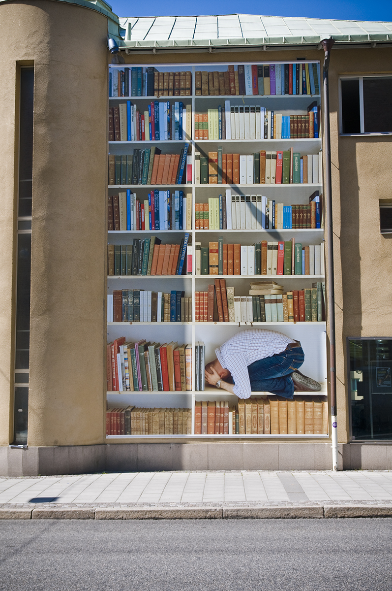 På en husfasad hänger en vepa av ett fotografi som föreställer en hög och överdimensionerad bokhylla och i ett av facket ligger en ihopkrupen man i rutig skjorta. 