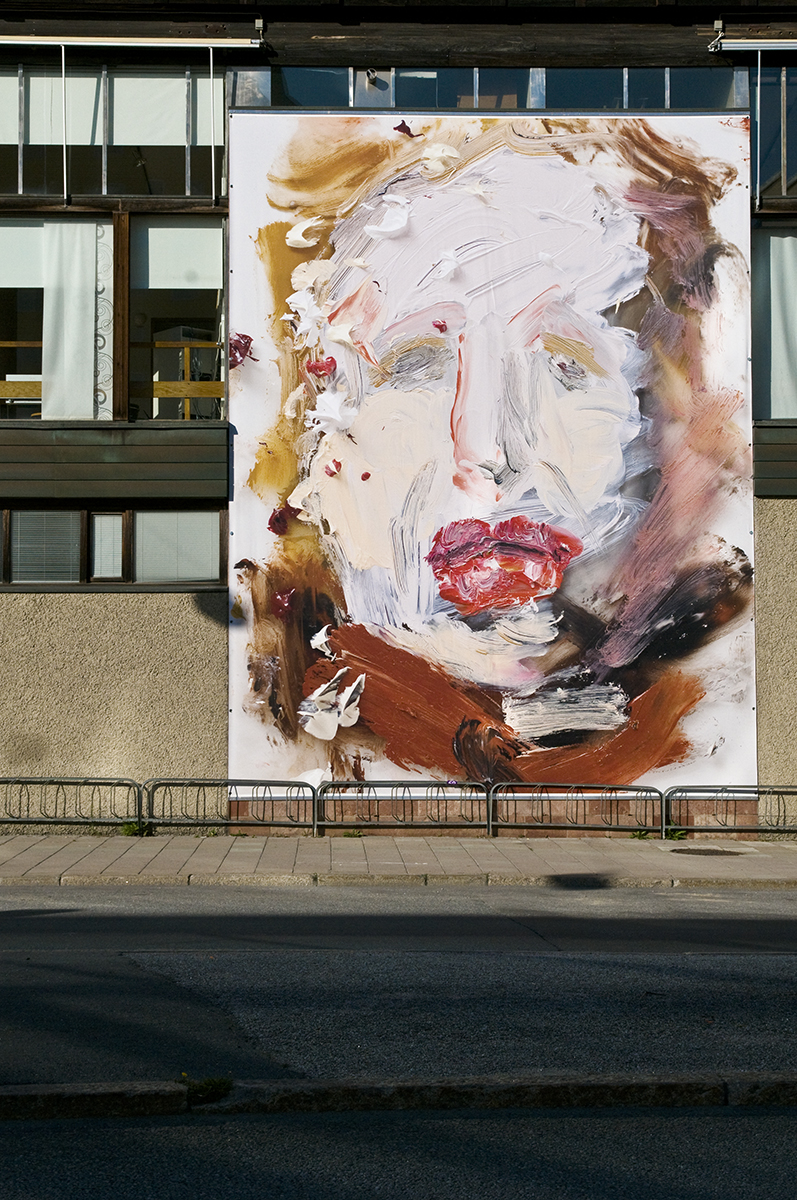 På en husfasad hänger en vepa med en abstrakt målning av ett ansikte. 