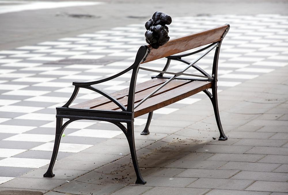 På en tom parkbänk på en gata balanserar ett svart objekt på ryggstödet. Objektet är en svart "klump" av ihoplindat tyg.