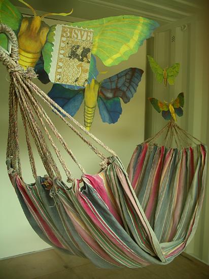 I ett litet rum hänger en randig hängmatta och runt i rummet flyger fyra stora fjärilar i olika storlekar och färger. 
