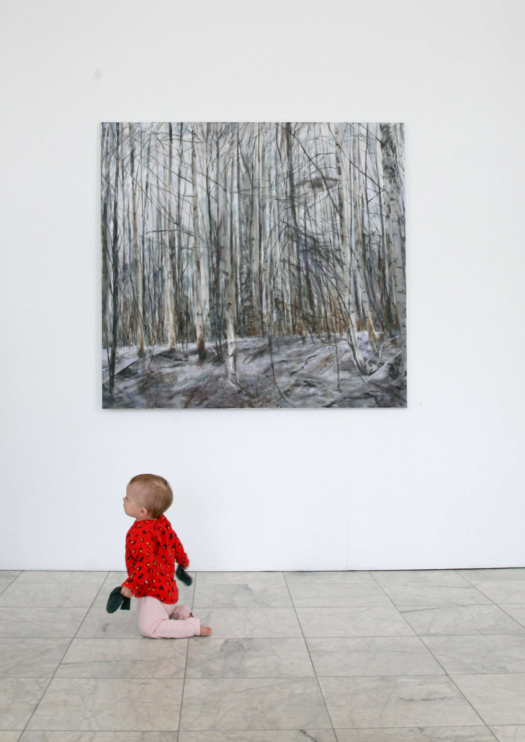 Ett barn kryper på golvet i konsthallen. På väggen hänger en tavla med motivet från en skog.