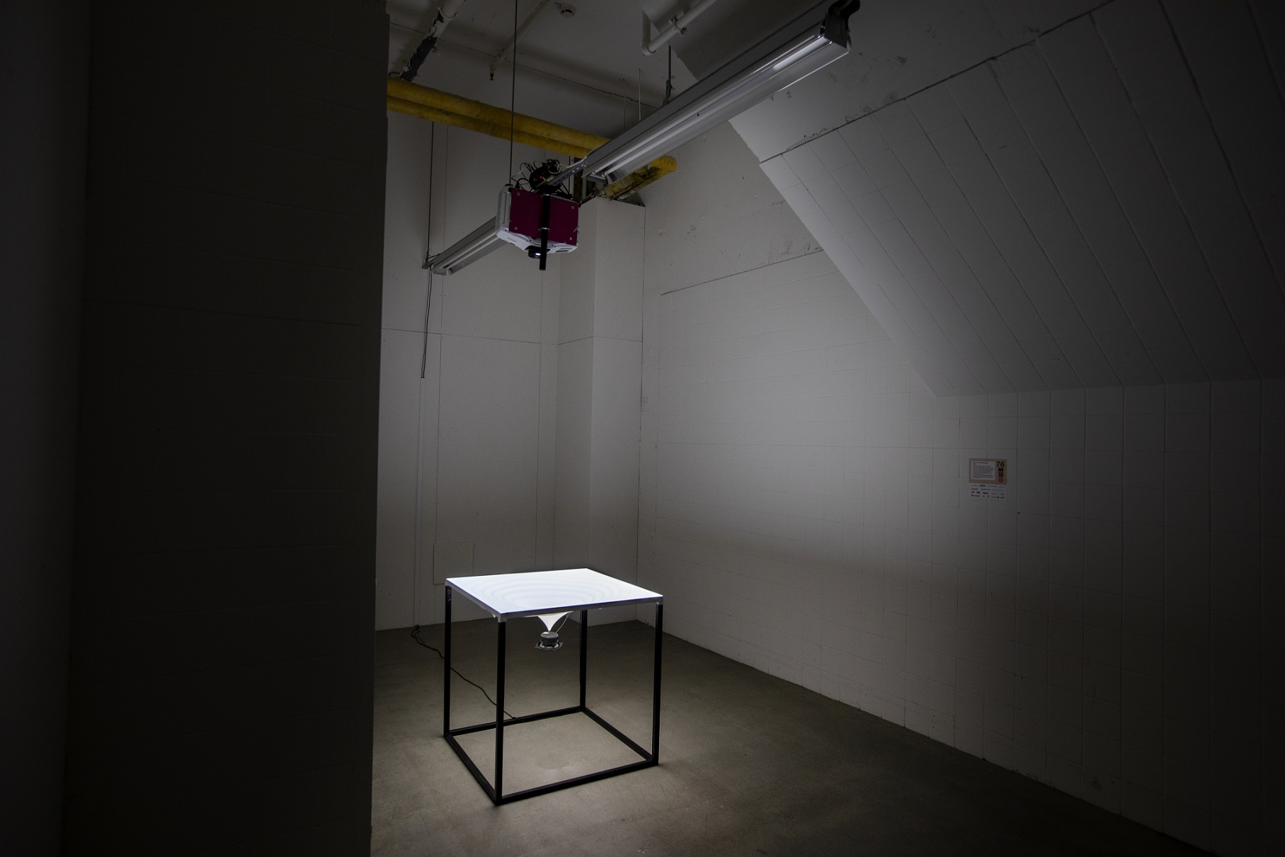 I hörnet av ett mörkt rum står ett vitt bord med ett spetsigt föremål under sig. I taket över hänger en projektor. 