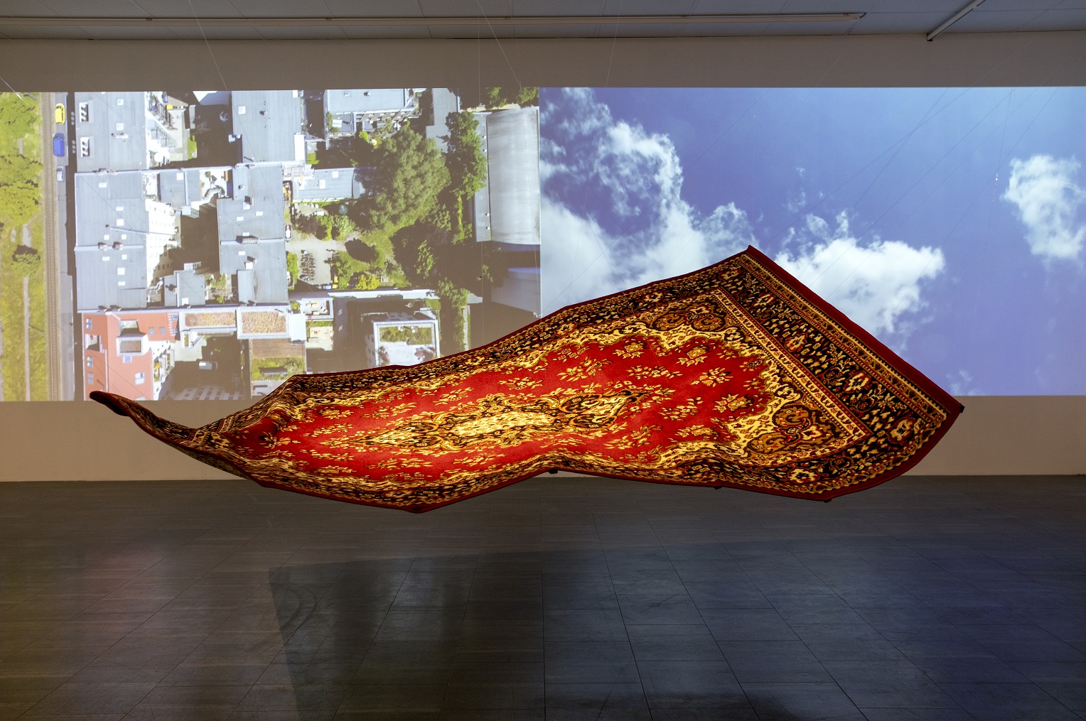 A red Persian carpet seems to float in the air in the middle of a room and in the background two photographs are projected onto a white wall. The image on the left is taken above a buildings and a road with cars and the second image is of a blue sky with clouds.