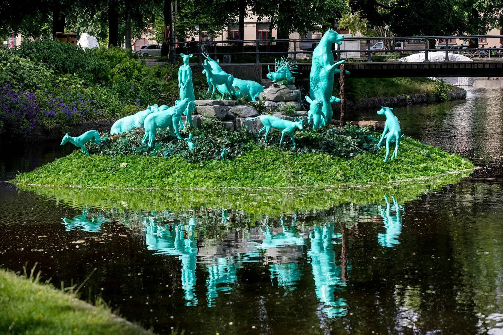 In the center of Örebro, a green island floats on the water surface of Svartån. The island has been occupied by 15 different wild animals depicted in green-blue lifelike statues. The island and the animals are reflected on the surface of the water.