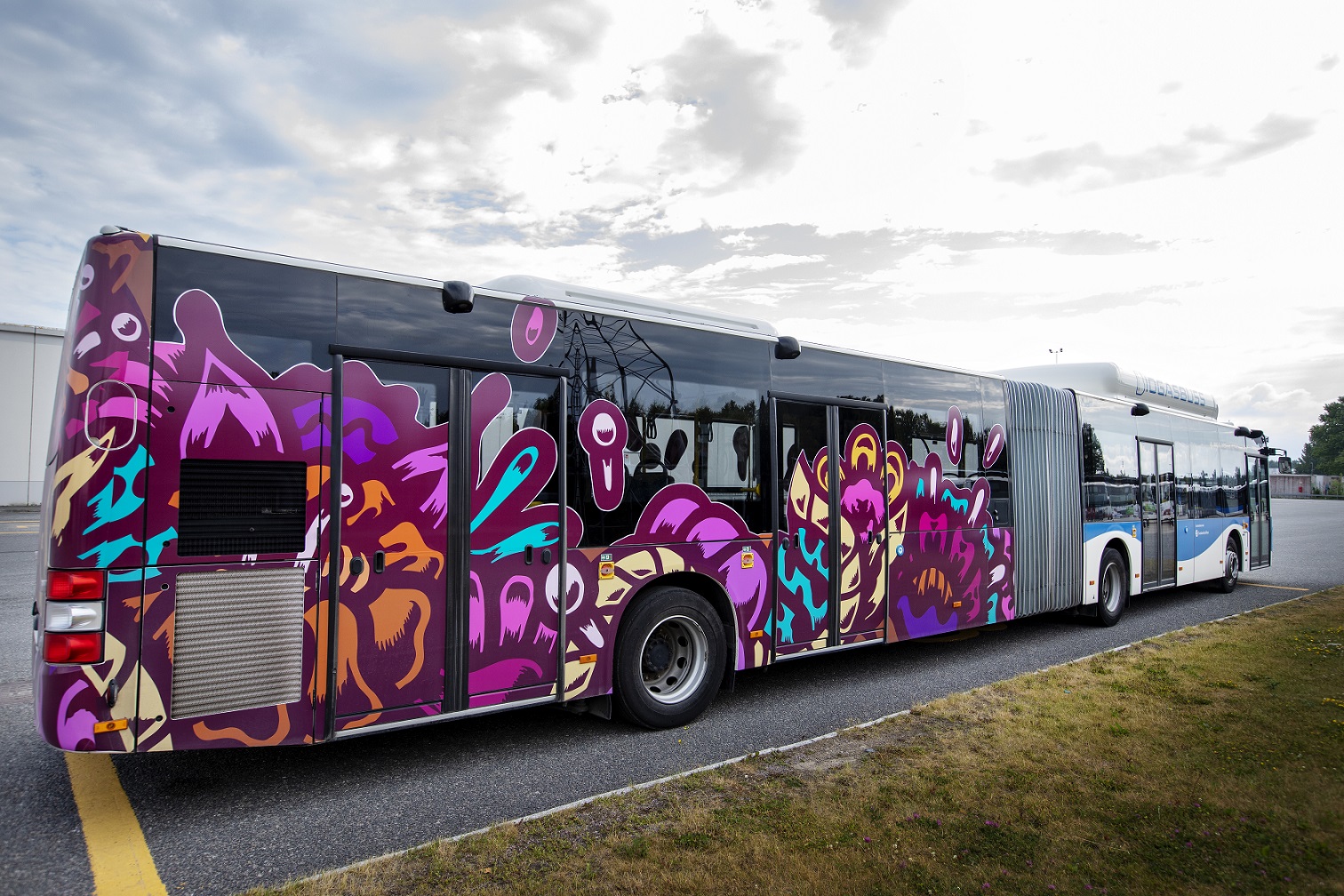 One of the city's city buses, an articulated bus has one half decorated with a print of a floral pattern. The pattern comes in different forms and is mainly in the color purple but also outlines in the colors yellow, orange, blue and pink.