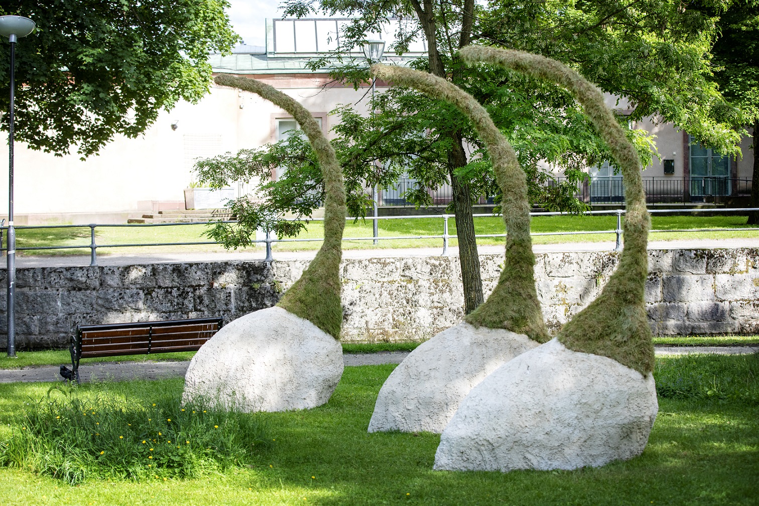 Tre konstverk växer upp ur gräsmattan. Underdelen består av en betong klump som sedan möter mossa som växer uppåt liknande en form som en tomteluva.