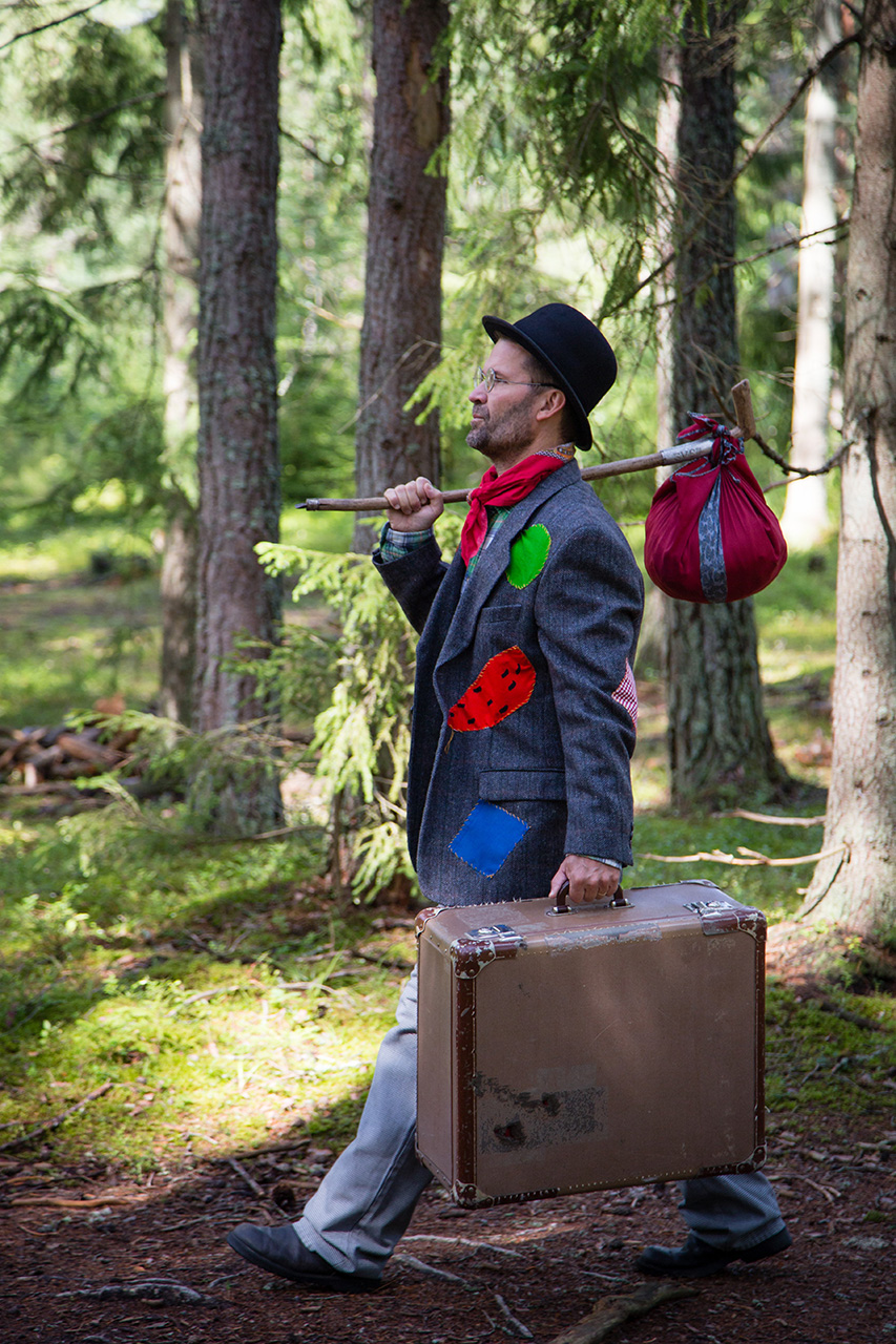 En man i slitna kläder går i skogen. Han bär en sliten väska och en matsäck på en pinne.