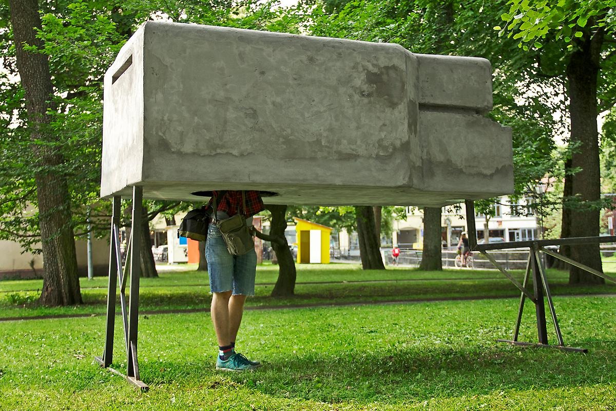 I en park på två stålställningar står en betongbunker med ett håll nertill så man kan stoppa in huvudet och kolla inuti bunkern. 