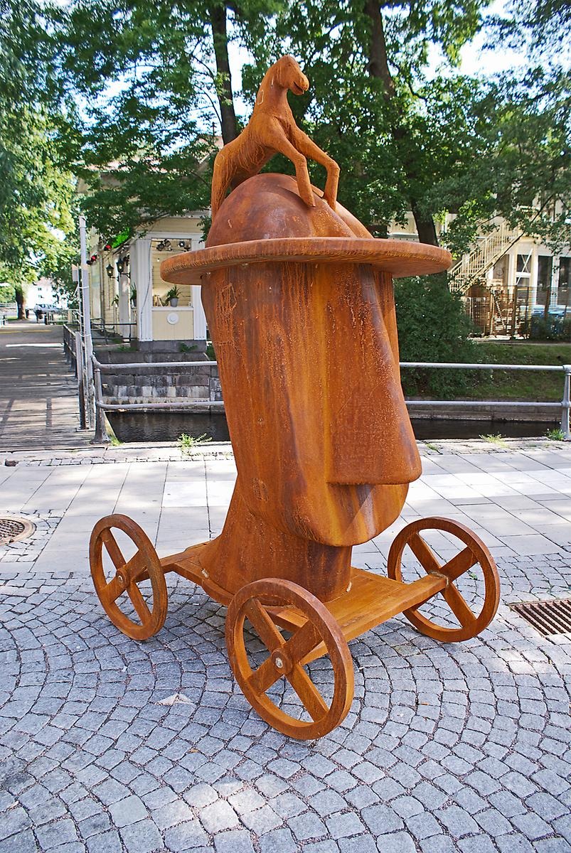 På kullerstenarna står en rostfärgad skulptur på en platt vagn med fyra hjul. På vagnen står ett stort människohuvud som har en hatt på sig samt på hatten står en liten häst. 