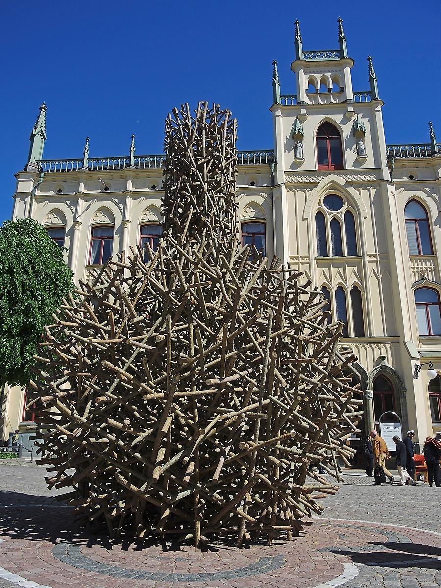 En solig dag i Örebro och framför Rådhuset står ett stort konstverk som genom massa tjocka träpinnar ihopsatta med varandra bildar en rund flaskform. 