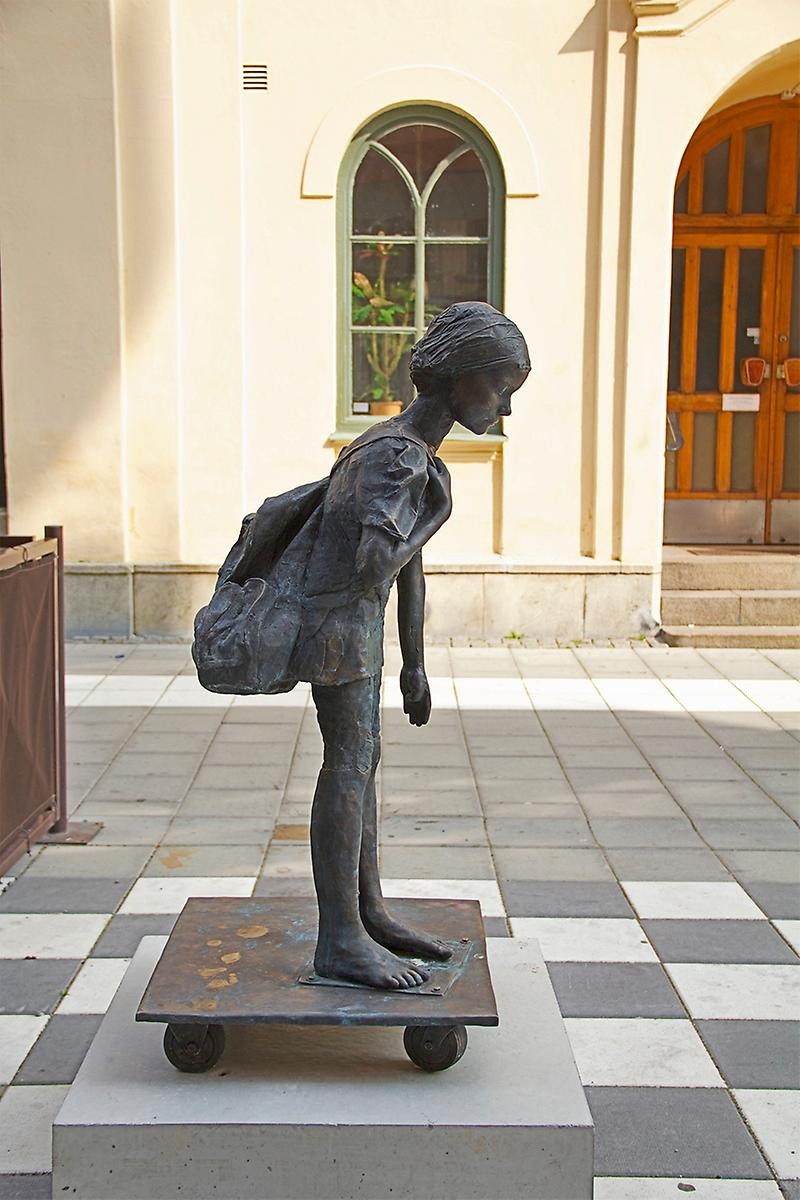 Utomhus på Kungsgatan står en barn skulptur i färgen grönbrun. Barnet bär på en ryggsäck och blundar med ett neråt sänkt huvud. På ena benet ser man en skarv då denna skulptur är avgjutningar från flera barn. 