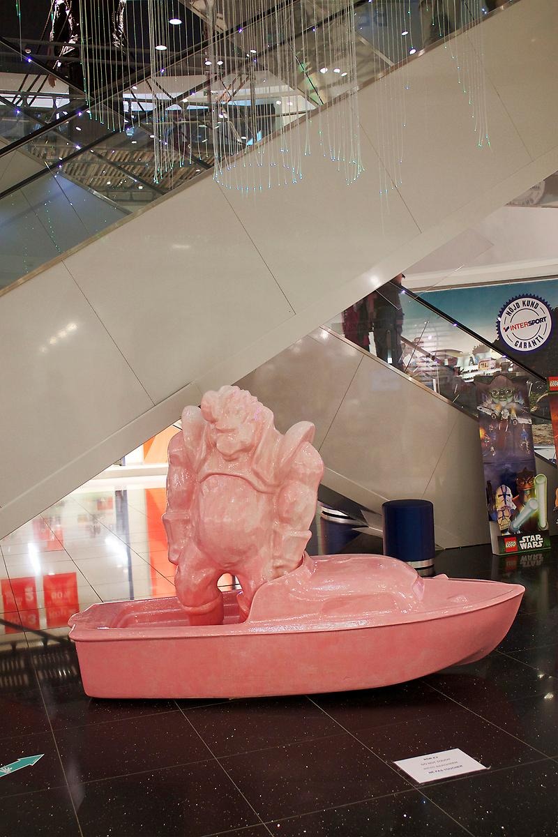 Inne i gallerian Vågen står en rosa skulptur framför rulltrappan. Skulpturen efterliknar en rosa monsterliknande man som står i en rosa båt. 