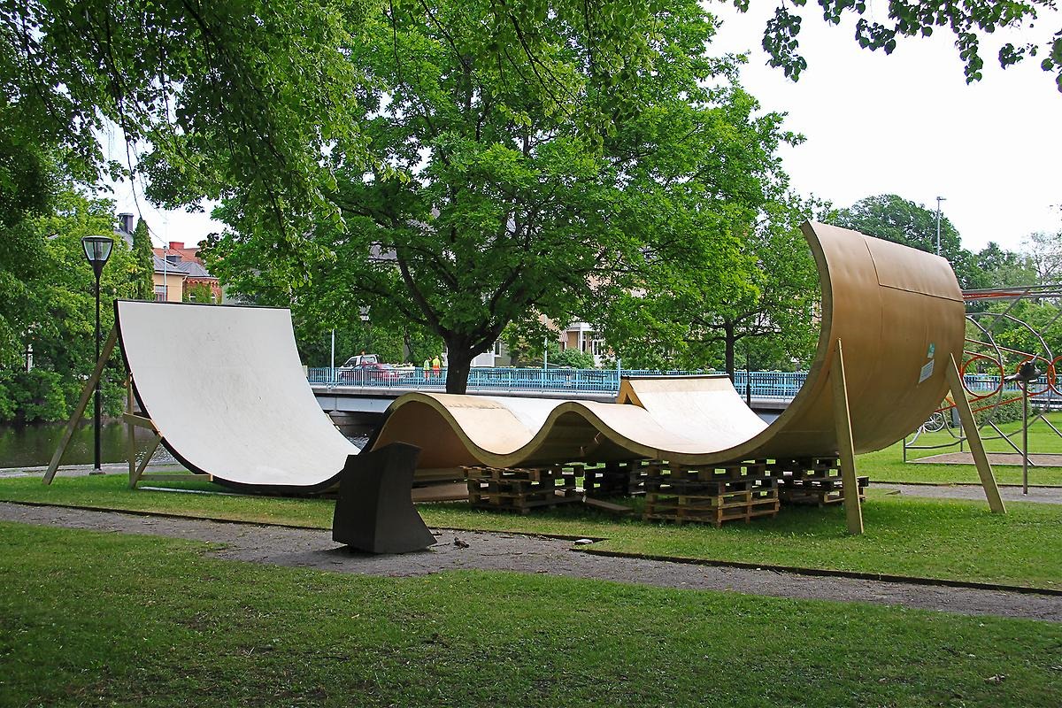 I en park står en stor skateboard ramp på gräsmattan. Rampen är byggd som rundade gupp lite som att åka över ett berg. 