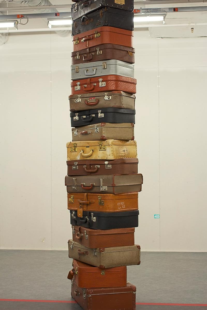 I ett vitt rum står 16 stycken kappsäckar staplade på varandra. Alla kappsäckarna har olika färger och läderaktigt material.