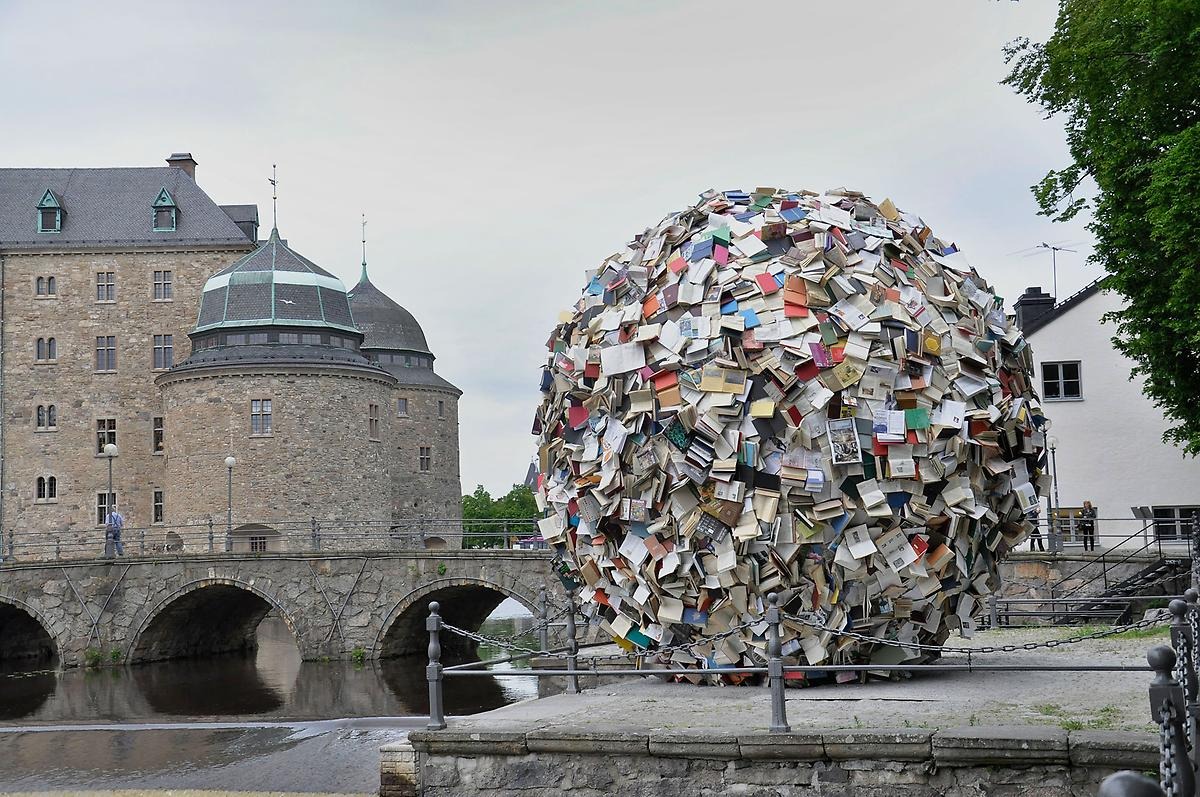 En stor rund skulptur utsmyckad med massa böcker som både är stängda och öppna. I bakgrunden skymtar man Örebro slott. 