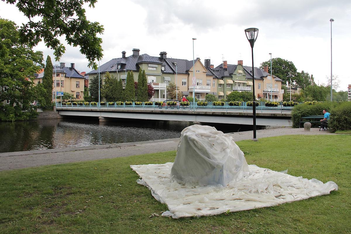 På en gräsmatta framför Svartån ligger en klump liknande en sten och över den har man lagt ett vit tyg i ett vågigt mönster. I bakgrunden ser man en kvinna som cyklar på den stor bron. 
