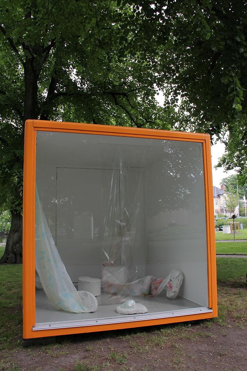 I en glasmonter med orangekanter ligger fem olika vita skulpturer och från två stycken går ett stycke plast upp mot taket. 