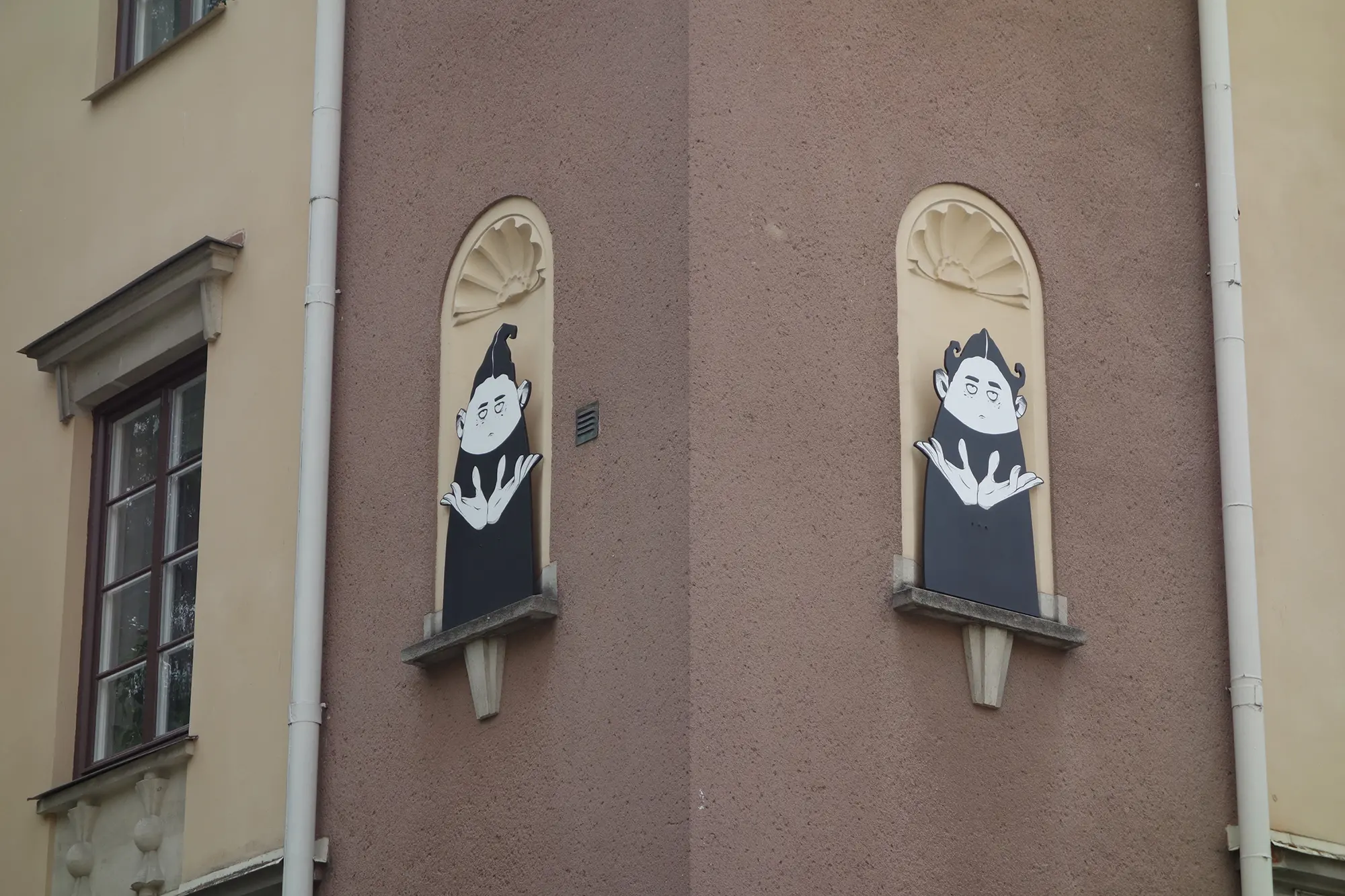 Högt upp på utsidan av en husfasad syns två stora illustrationer i seriestil föreställande figurer i svartvitt på var sin vägg. Deras ansiktsuttryck är neutrala och de har stora händer riktade uppåt.