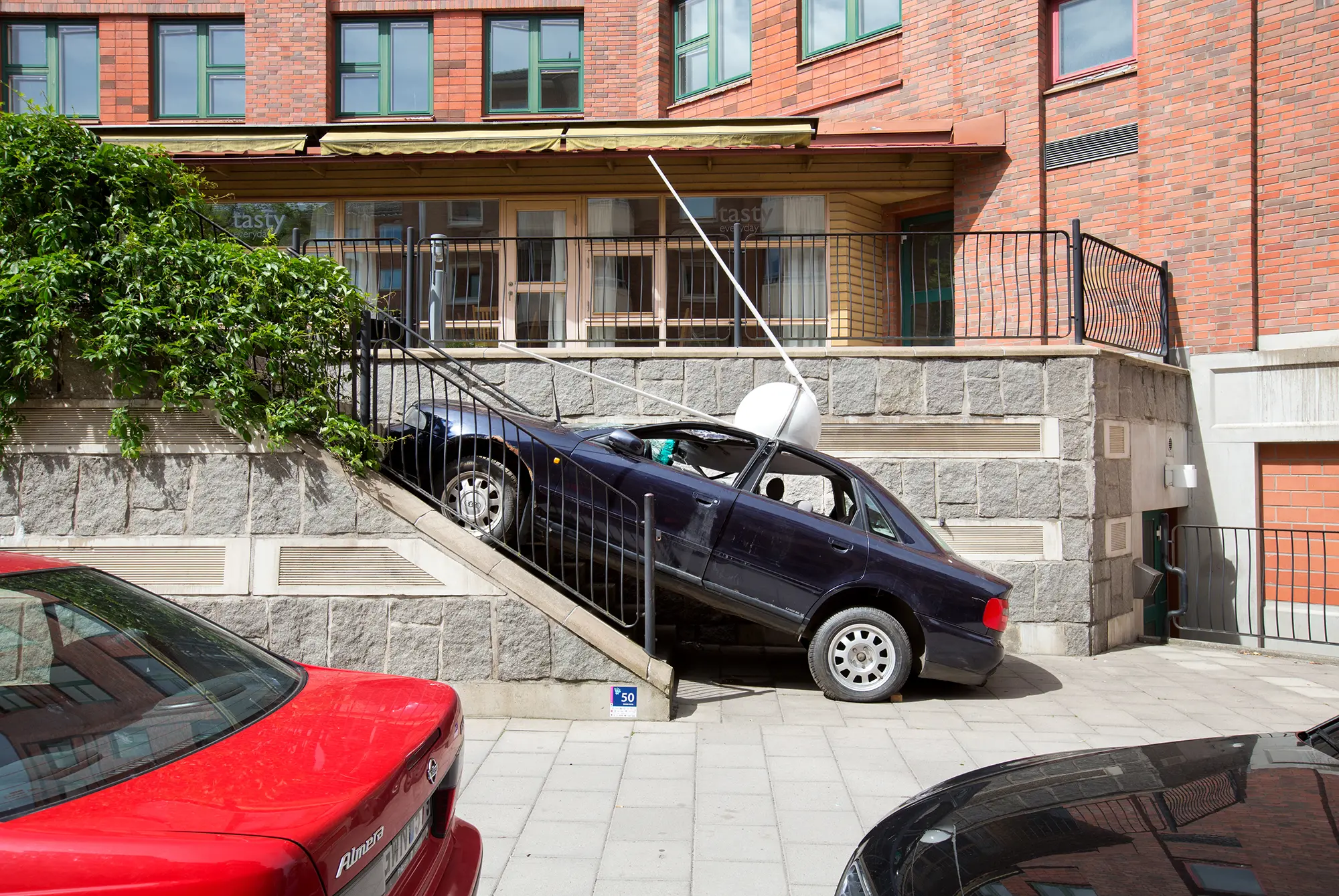 En sliten mörkblå bil står uppställd lutande på en trappa. Framhjulen står på trappan och bakhjulen står på marken. På bilens tak finns en vit klotformad skulptur med två vita pinnar vinklade framåt.