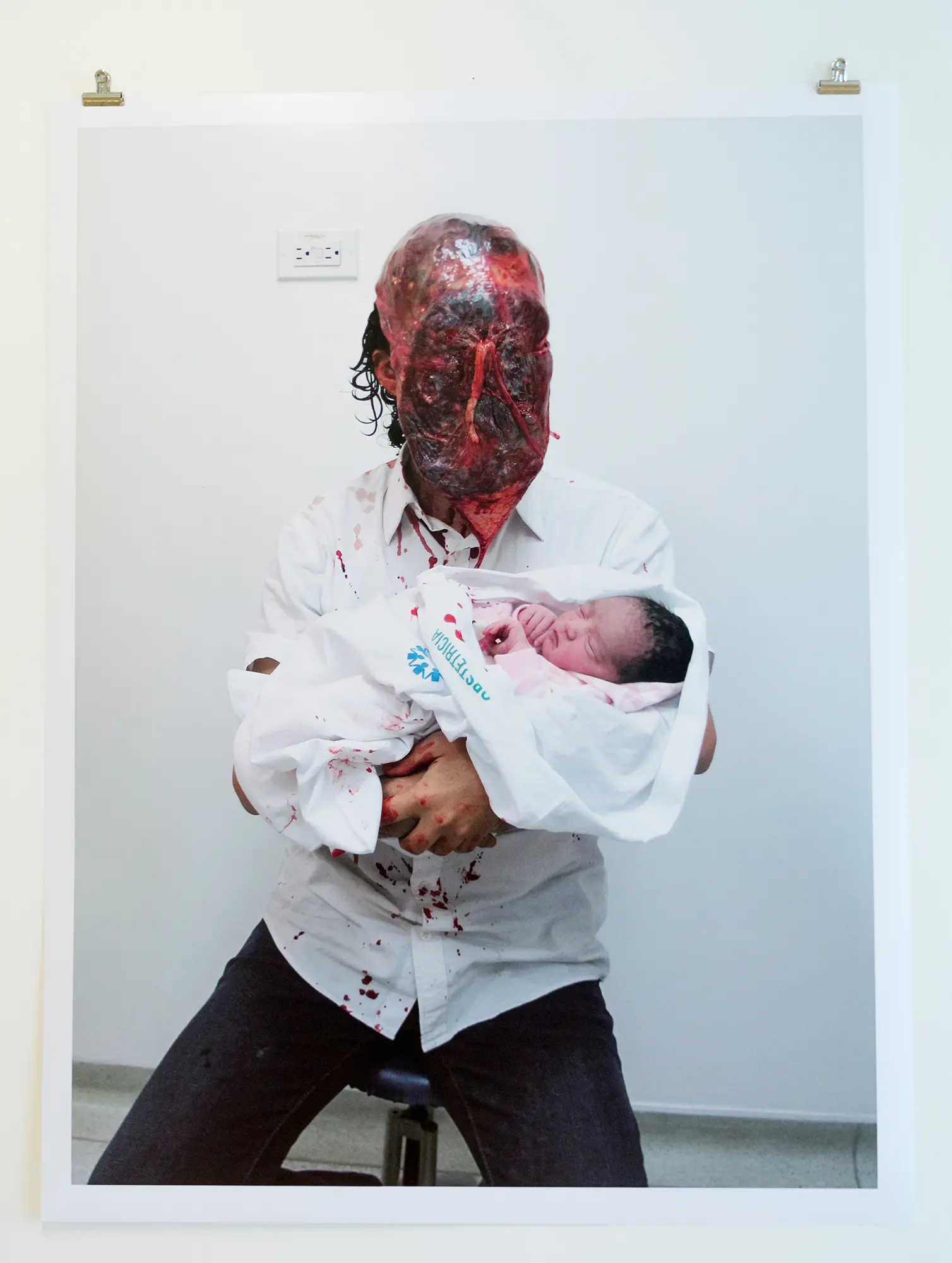 På en upphängd fotoplansch syns sittande en manlig figur. Figuren bär en mask som gör att det ser ut han som att han har ett stort sår över hela ansiktet. På hans skjorta syns röda fläckar. I sin famn håller han ett litet barn.