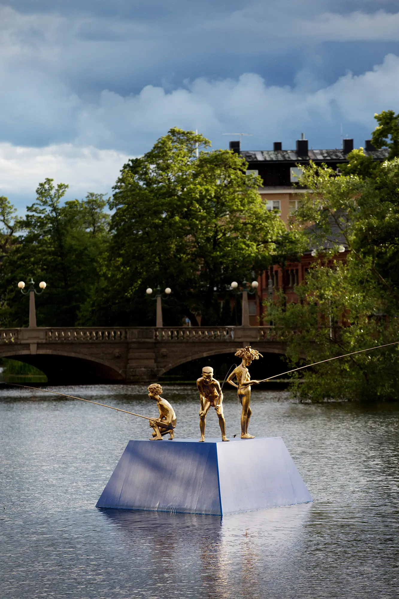 På en blå plattform ute i en å står tre guldfärgade statyer föreställande tre pojkar hållandes fiskespön. Två av pojkarna står framåtlutade och en står lätt bakåtlutad. I bakgrunden syns en bro och gröna träd.